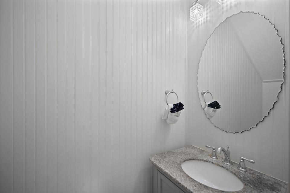 Chic half bathroom with white beadboard walls and a granite countertop. Features sleek chrome fixtures and a modern light fixture for a clean, coastal look.