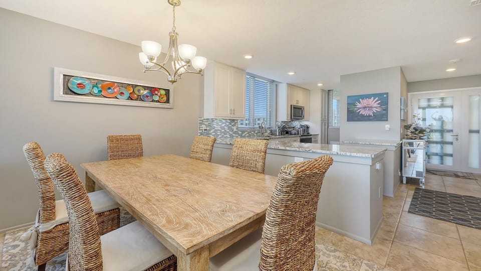 Woven dining chairs surround a light wood table beneath a brushed nickel chandelier and colorful wall art.