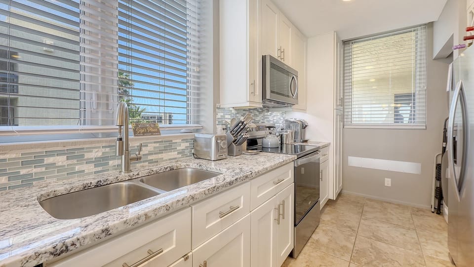 Galley kitchen with granite countertops, glass tile backsplash, stainless steel appliances, and large windows.