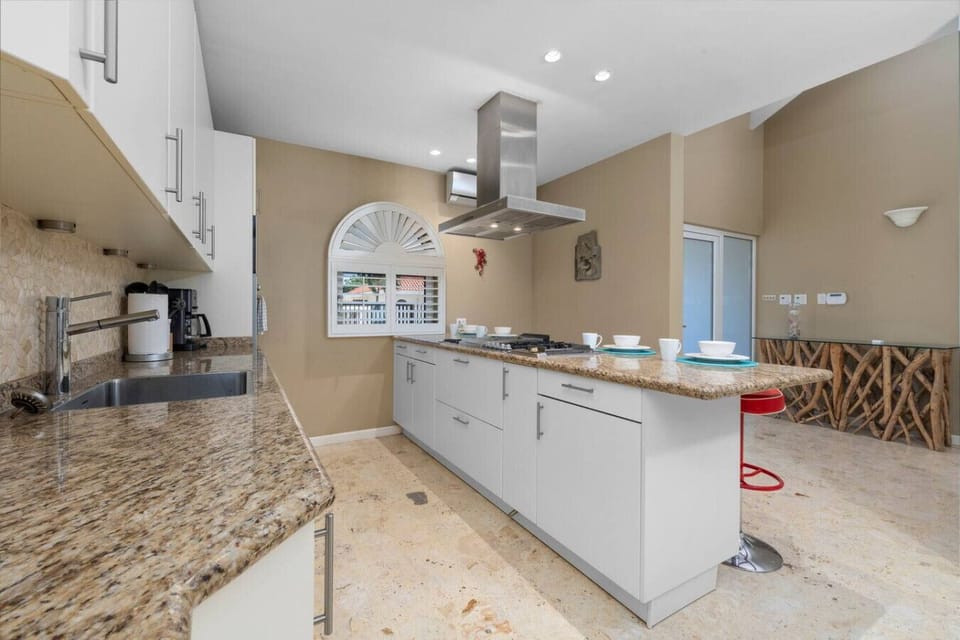 This modern kitchen offers white cabinetry, granite countertops, stainless steel appliances, and a cozy breakfast bar with red stools, complemented by natural light from an arched window.