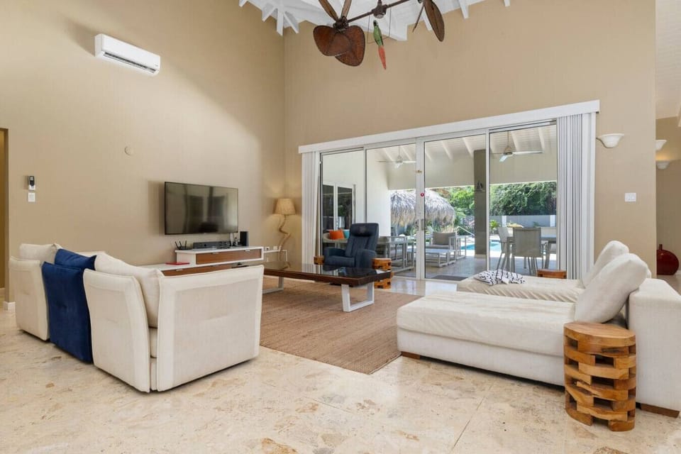 This bright living room offers comfy seating, a sleek coffee table, and a large TV. Glass doors open to a poolside patio, blending indoor and outdoor living in a spacious, high-ceilinged space.