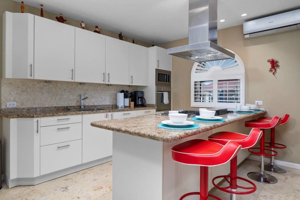 This modern kitchen offers white cabinetry, granite countertops, and high-end appliances, with a stylish breakfast bar and red stools for casual dining.
