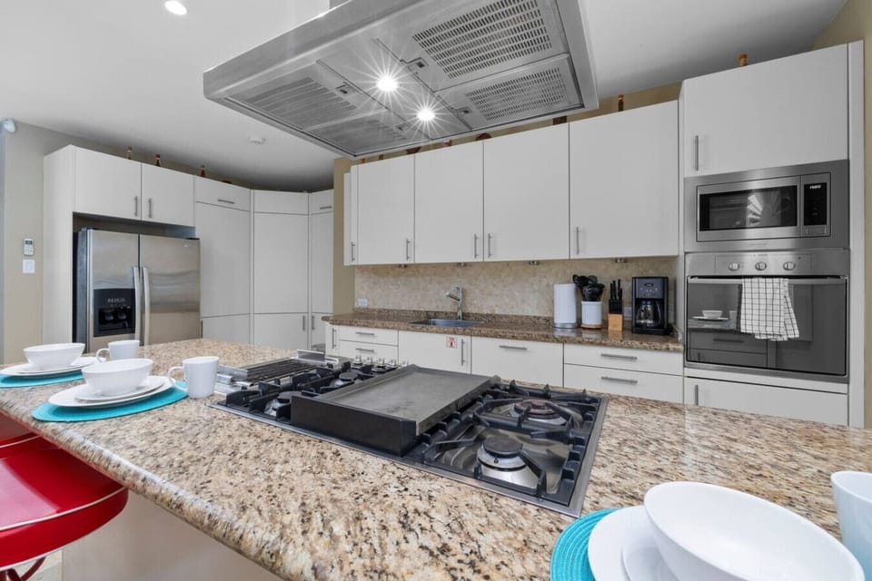 This modern kitchen features sleek white cabinetry, granite countertops, and high-end stainless steel appliances, including a gas stove, microwave, and fridge. The stylish breakfast bar with red stools is perfect for casual dining or morning coffee.