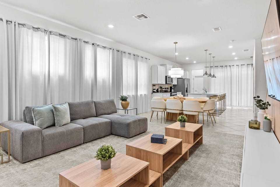 Relax in the spacious living area, where a contemporary gray sofa with soft cushions awaits you. Large windows fill the room with natural light, showcasing the sleek wooden furniture and stylish decor for a welcoming atmosphere.