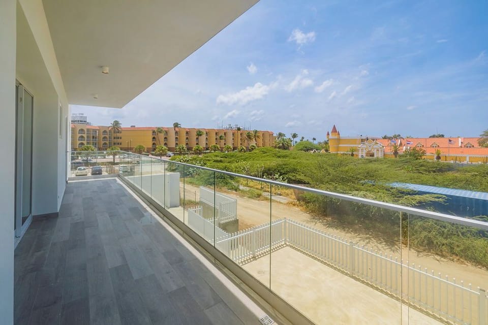 A spacious balcony with glass railings overlooks the surrounding area, featuring lush greenery and nearby colorful resort-style buildings under a clear blue sky.