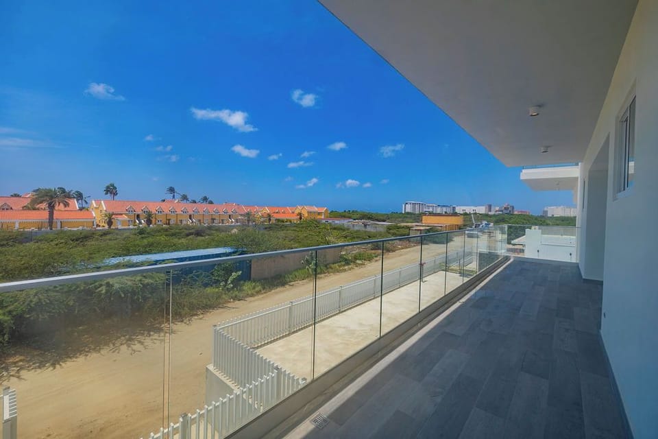 The property features an expansive modern balcony with glass railings and views of nearby residential neighborhoods, natural dunes, and coastal areas.