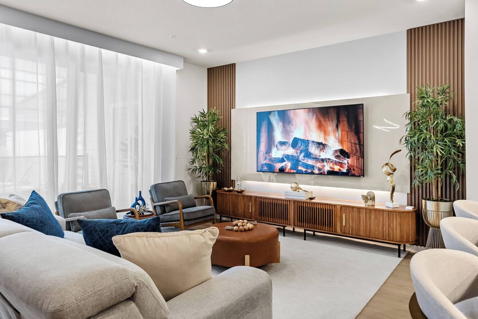 Unwind in the cozy living room with plush gray seating and soft blue accents. Enjoy movie nights on the large TV framed by elegant wood paneling, all while natural light gently filters through sheer curtains.