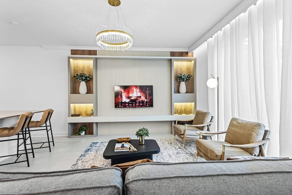 Welcome to a bright living area filled with natural light, featuring a soft gray sofa and chic gold-accented armchairs. Enjoy the modern design with a sleek wall-mounted TV and a cozy fireplace, perfect for relaxation.