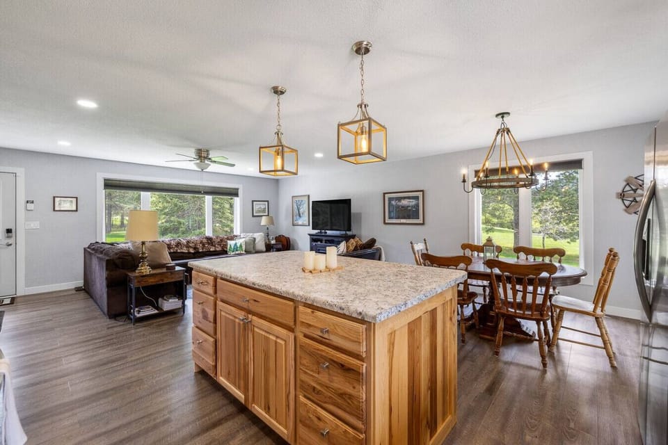 This bright, open-plan interior features a central wooden kitchen island, seamlessly connecting to a cozy living room with a large window and a dining area, creating a spacious and inviting common living space.