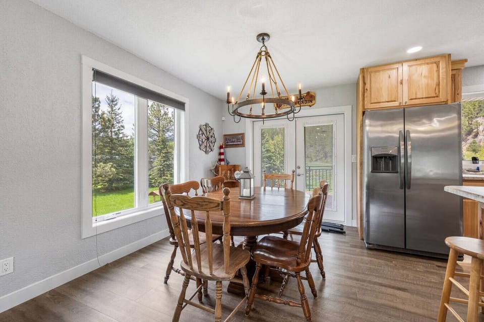 Gather around the rustic dining table in this light-filled space, where large windows frame peaceful forest views and a charming chandelier adds a cozy cabin touch.
