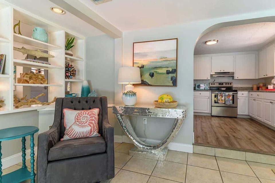 Bright dining area with large windows and sliding doors to the patio. Includes built-in shelving, cozy armchair, modern kitchen with stainless steel appliances.