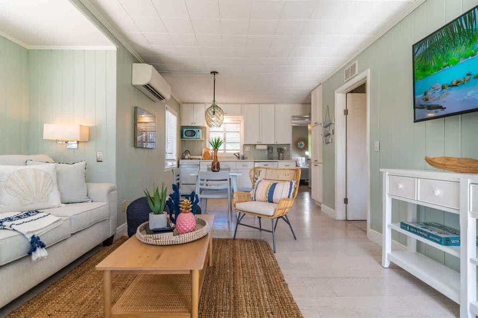 Living space features a plush sofa, accent chairs, TV, and a coffee table. The open layout connects to a kitchen with full appliances, dining table, and stylish pendant lighting.