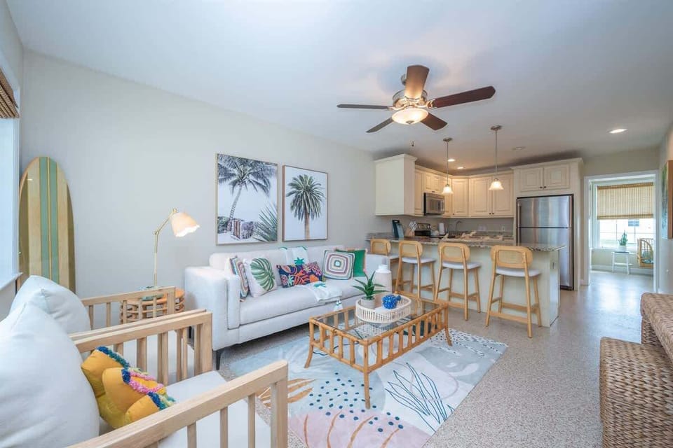 Cheerful living space with a sofa, accent chairs, wicker coffee table, and bar seating. Features coastal artwork, colorful pillows, a smart TV, and a ceiling fan—all giving a beach-inspired setting.