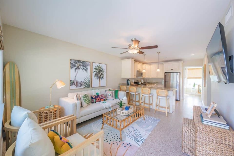 Cheerful living space with a sofa, accent chairs, wicker coffee table, and bar seating. Features coastal artwork, colorful pillows, a smart TV, and a ceiling fan—all giving a beach-inspired setting.