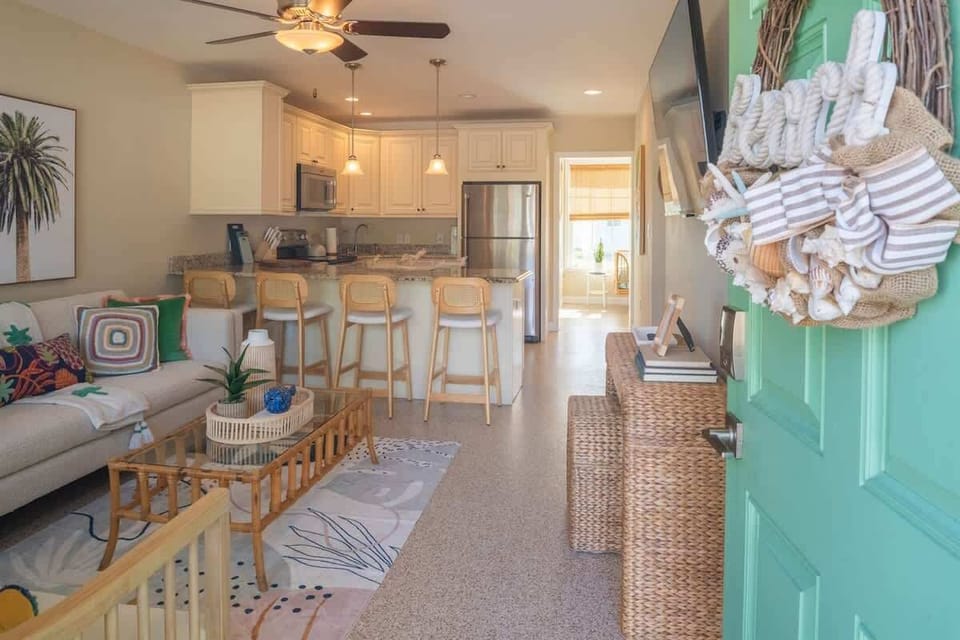 Cheerful living space with a sofa, accent chairs, wicker coffee table, and bar seating. Features coastal artwork, colorful pillows, a smart TV, and a ceiling fan—all giving a beach-inspired setting.