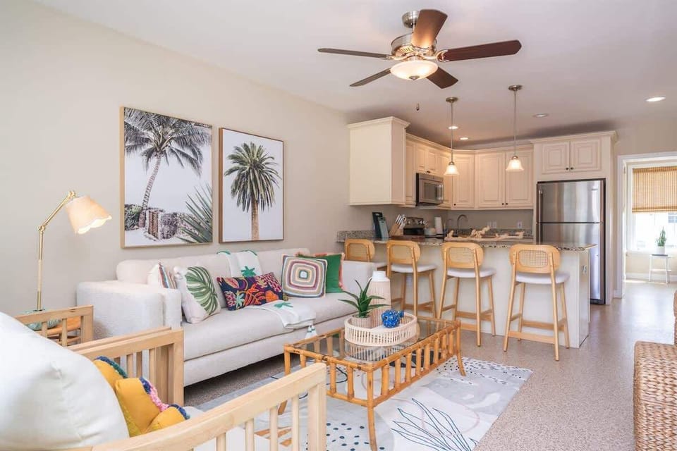 Cheerful living space with a sofa, accent chairs, wicker coffee table, and bar seating. Features coastal artwork, colorful pillows, a smart TV, and a ceiling fan—all giving a beach-inspired setting.