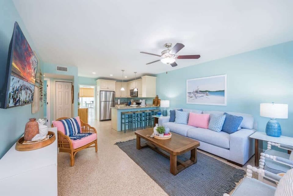 Open-concept living room with a plush sofa, accent chairs, a smart TV, and a wood coffee table. Coastal decor, pastel tones, and woven accents complete the relaxed vibe.