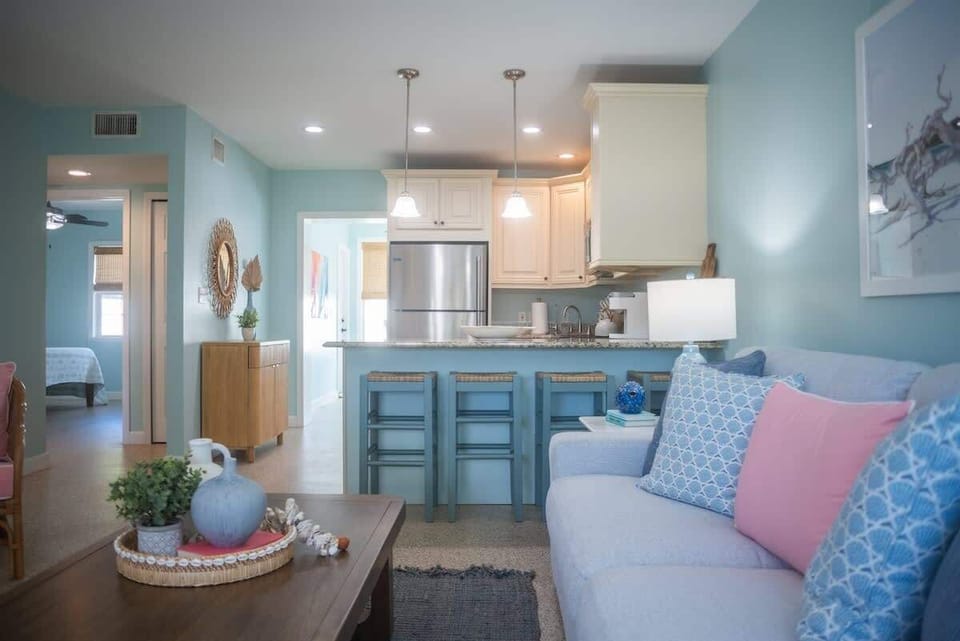 Open-concept living room with a plush sofa, accent chairs, a smart TV, and a wood coffee table. Coastal decor, pastel tones, and woven accents complete the relaxed vibe.