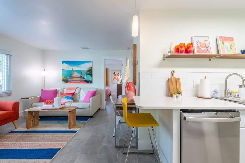 Living area with a leather sofa, bright accent chairs, and a wall-mounted TV. Includes a dining nook, workspace, and open kitchen.