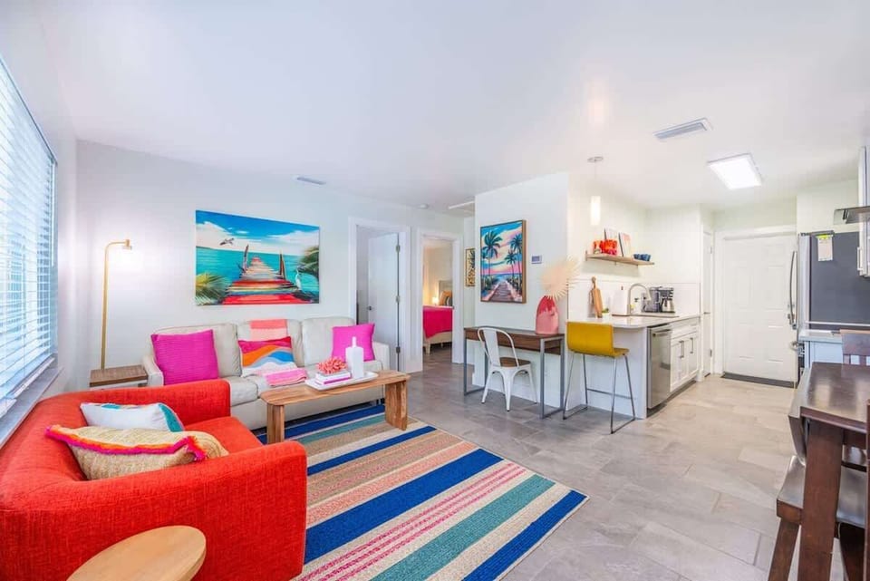 Living area with a leather sofa, bright accent chairs, and a wall-mounted TV. Includes a dining nook, workspace, and open kitchen.