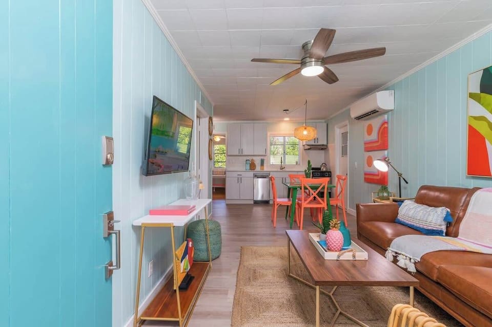 Tropical living room with leather seating, rattan accents, a smart TV, ceiling fan, bright decor, and open layout to the kitchen and dining area.