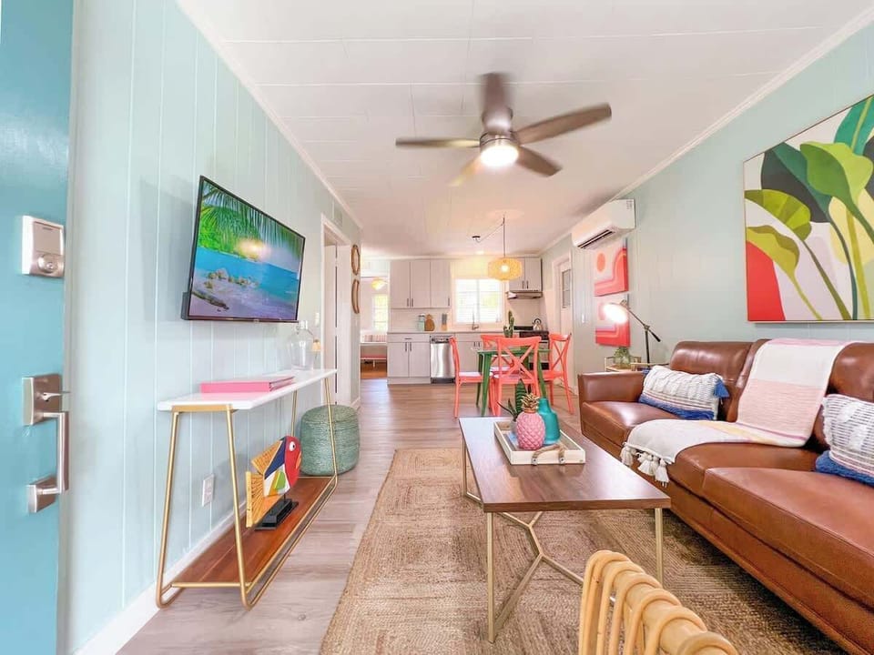Tropical living room with leather seating, rattan accents, a smart TV, ceiling fan, bright decor, and open layout to the kitchen and dining area.