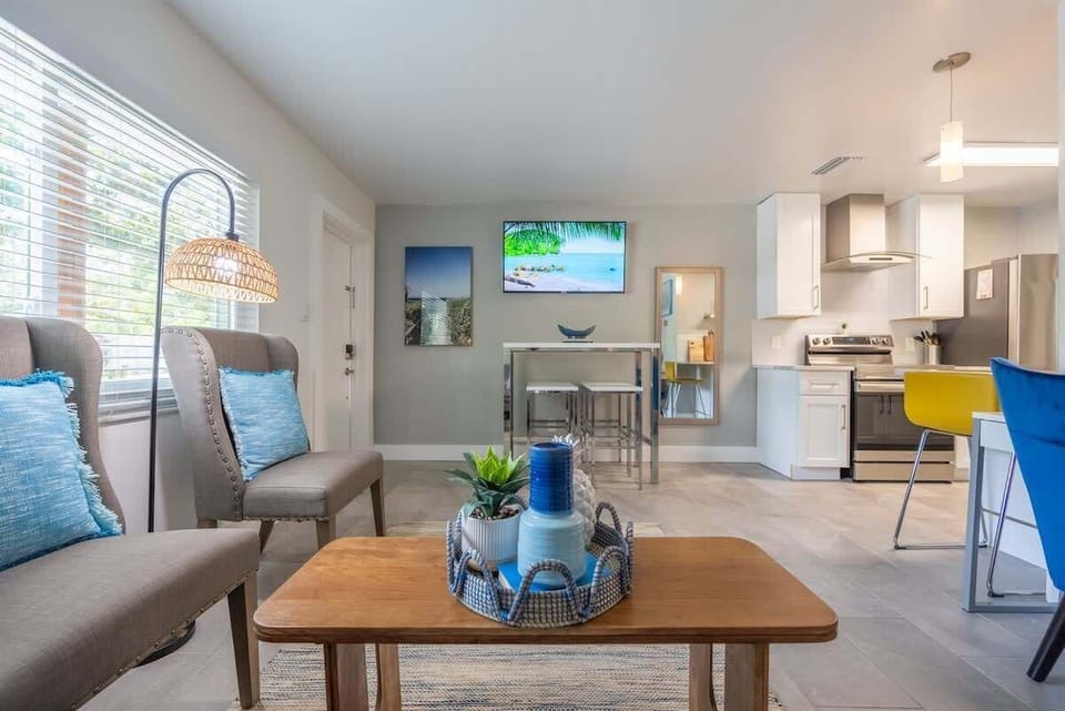 Open-plan living space with a full kitchen, breakfast bar, and dedicated workspace. Features modern appliances, comfy seating, and beachy decor throughout. Includes a mounted TV, dining nook, and vibrant accent furniture.