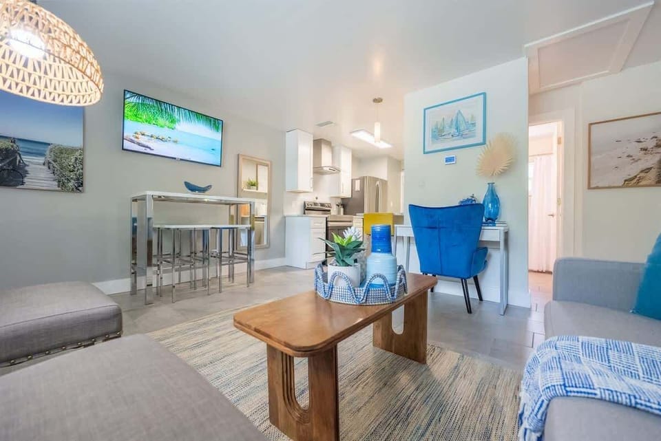 Open-plan living space with a full kitchen, breakfast bar, and dedicated workspace. Features modern appliances, comfy seating, and beachy decor throughout. Includes a mounted TV, dining nook, and vibrant accent furniture.