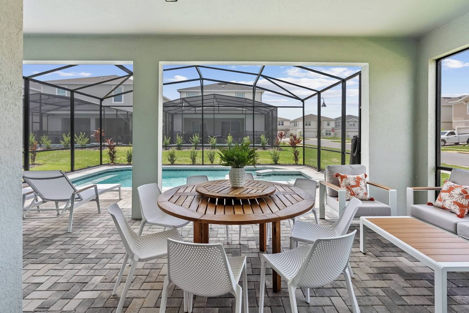Experience the charm of our outdoor dining area with a round wooden table and breezy white chairs, where you can enjoy lively meals while taking in views of the lush lawn and sparkling pool, ideal for relaxation.