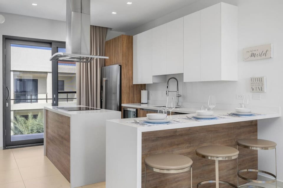 A contemporary kitchen with a bright, airy feel. White cabinetry paired with wooden accents and modern appliances creates a sleek, functional cooking space, complete with a breakfast bar and minimalistic design.