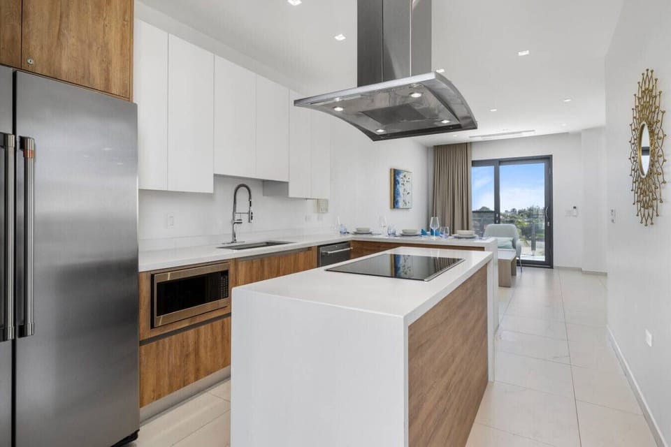 A contemporary kitchen featuring a spacious central island with a built-in cooktop. Stainless steel appliances, including a refrigerator and microwave, complement the bright, open space, with large windows offering a view of the outdoors.