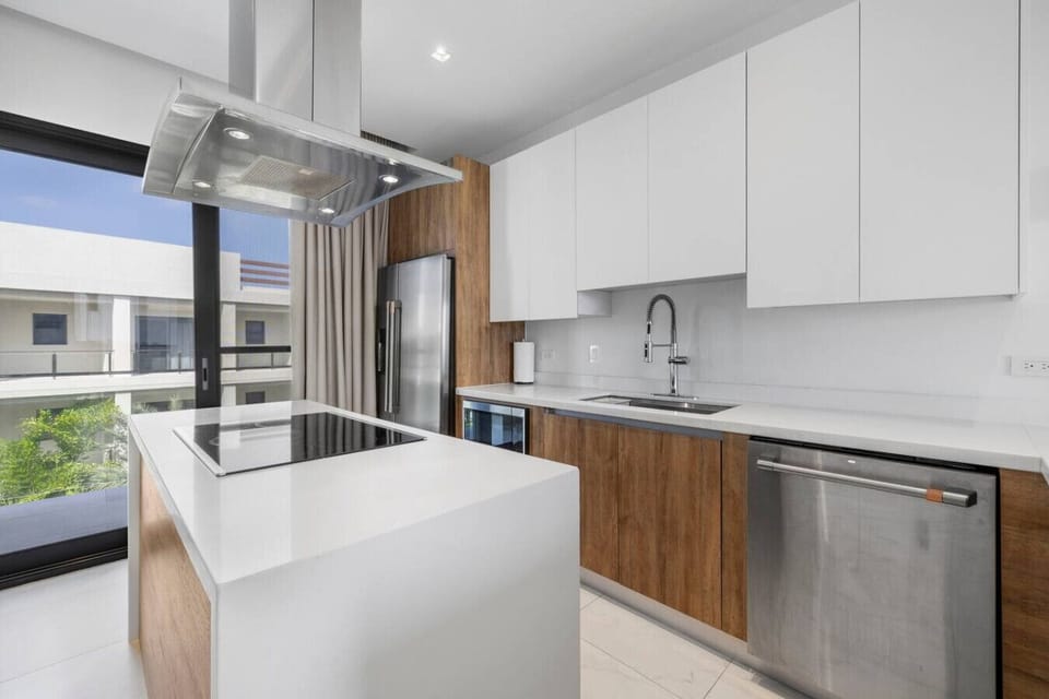 A contemporary kitchen with sleek white countertops and wooden accents. The kitchen features a large island, modern appliances including a dishwasher, and a window offering a view of the outdoors.