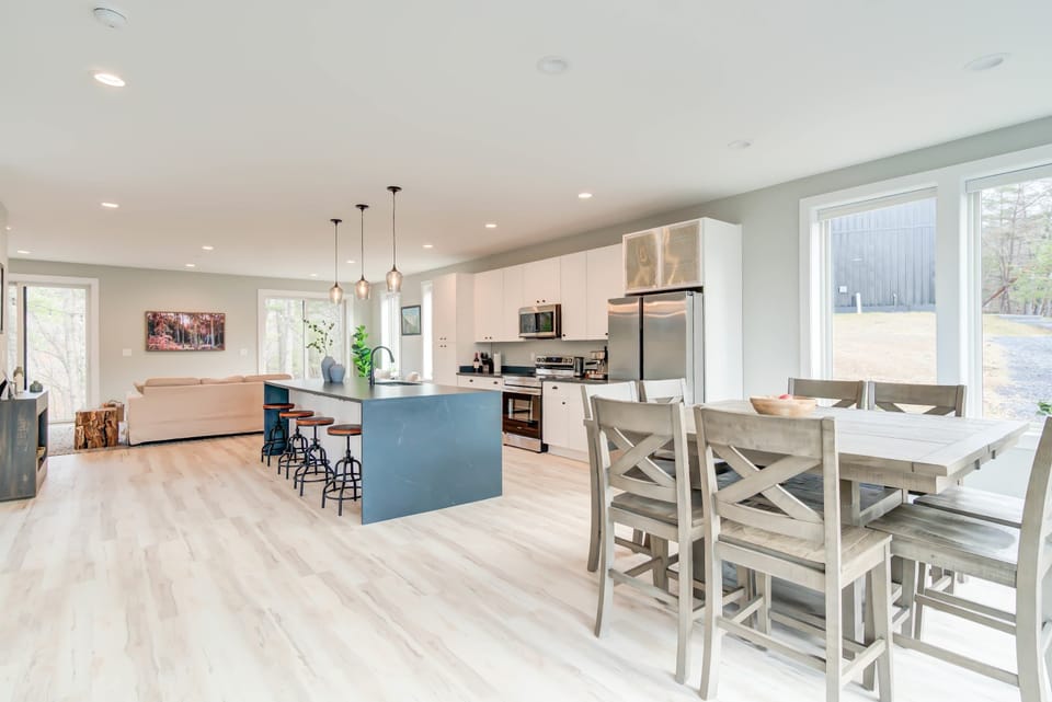An open, bright space featuring a kitchen with an island table, living room, and dining set, all framed by stunning views of nature through the glass windows.