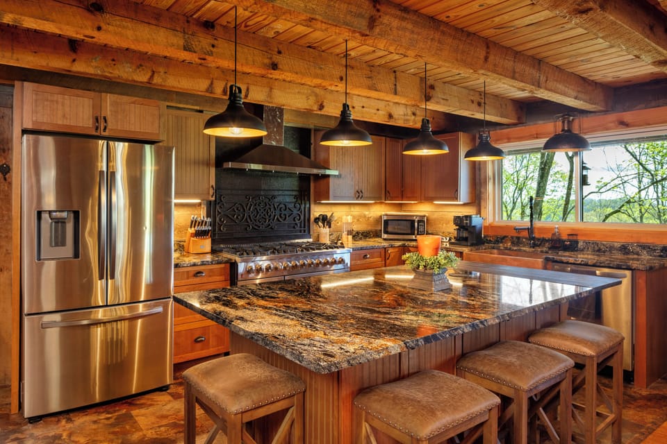 Gourmet mountain kitchen with granite counters, pro-grade gas range, stainless steel appliances & custom cabinets. Gather at the island for coffee or unwind with forest views through the large window—rustic charm meets modern comfort.