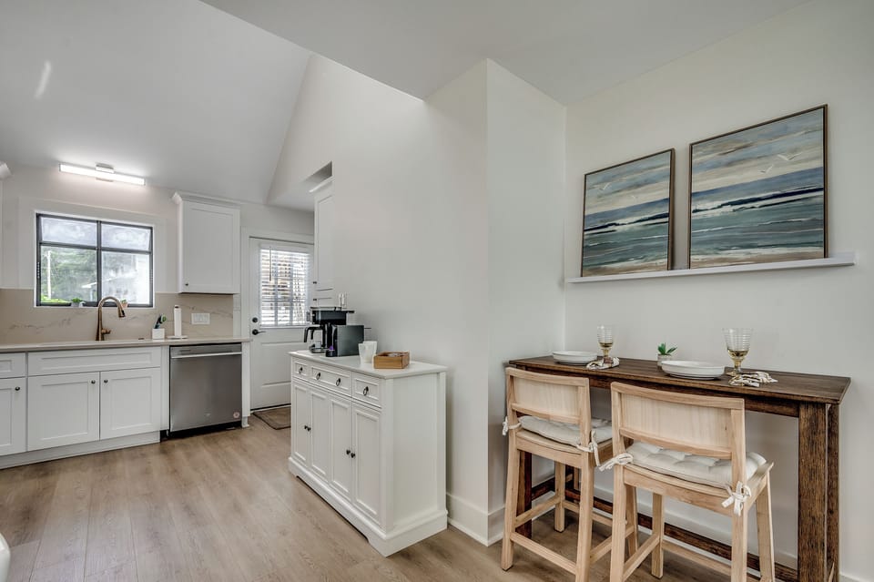 Small space, big charm! Enjoy a cozy bite at this stylish dining nook with breezy beach art and a peek into the dreamy white kitchen.