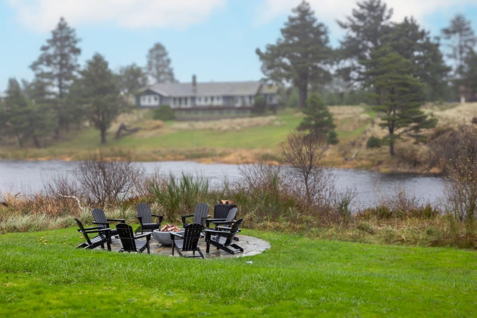 A fire pit and Adirondack chairs sit on a lawn overlooking water and distant trees.