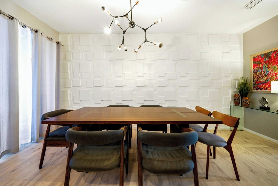 Sit at our stunning wooden dining table surrounded by cozy mid-century chairs. Enjoy the natural light streaming through elegant curtains, which enhance the textured white wall and bring warmth to the vibrant artwork.