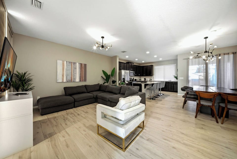 Relax in a bright, spacious living area with a sleek charcoal L-shaped sofa and modern light fixtures. The open layout seamlessly connects to a stylish dining space with a wooden table, ideal for your gatherings.