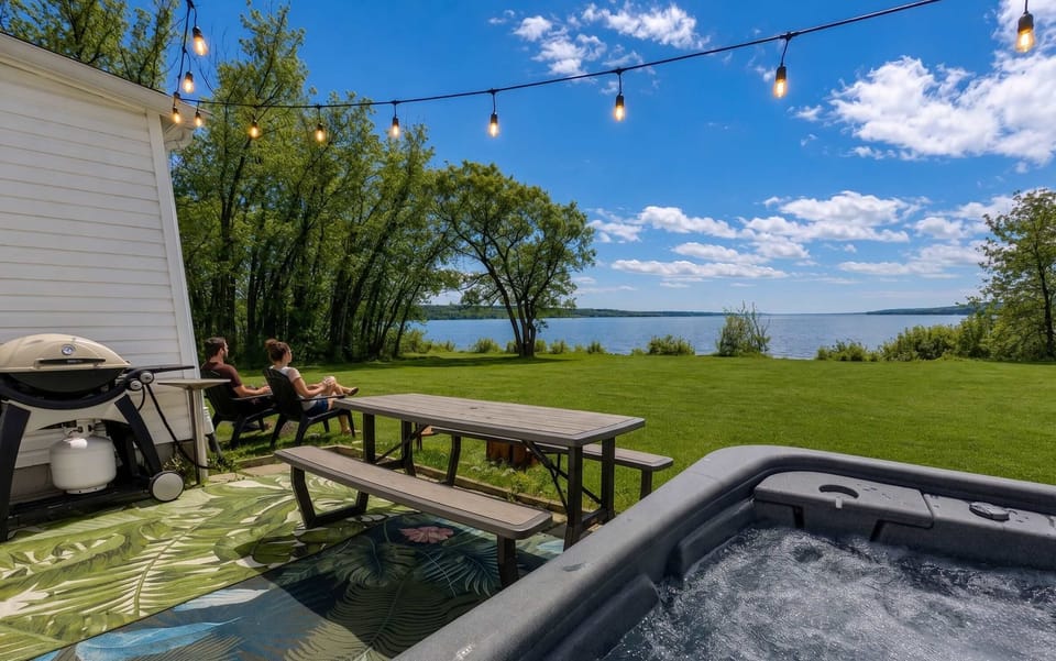 Grand terrain au bord de l'eau comprenant un bain tourbillon, un coin repas avec table de pique-nique, un barbecue et de charmantes guirlandes lumineuses.
