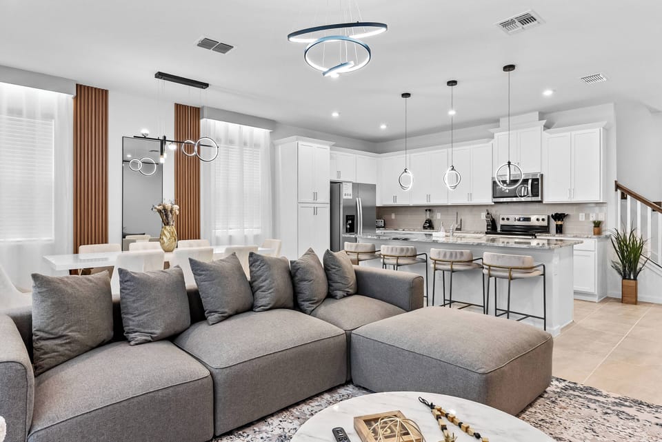Welcome to this inviting living area, filled with natural light pouring through sheer drapes. The plush gray sectional, chic coffee table, and modern kitchen create a stylish and comfortable space perfect for gatherings.