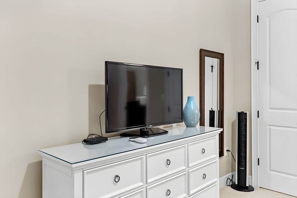 Relax in this cozy bedroom featuring a sleek white dresser topped with a flat-screen TV, perfect for movie nights. A striking blue vase adds a pop of color, while warm neutral walls create a calm and inviting atmosphere.