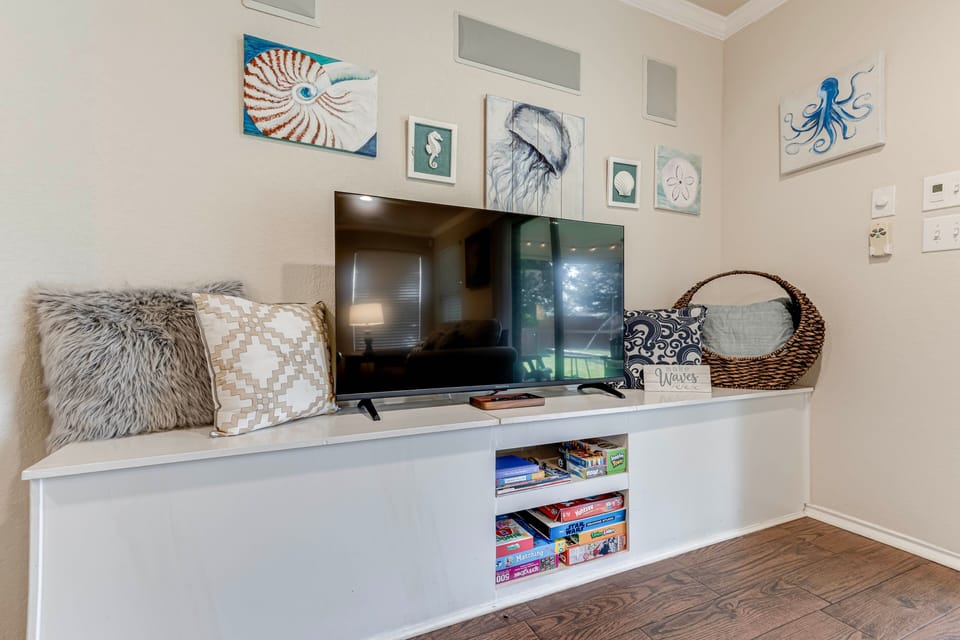 A flat-screen TV rests on a low console with games, flanked by coastal artwork.