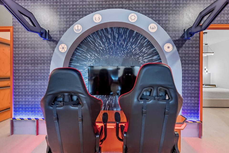 Experience a unique gaming zone with sleek black racing-style chairs in front of a large screen, all set against a striking futuristic backdrop. The ambient blue lighting enhances the excitement for an immersive gaming experience.