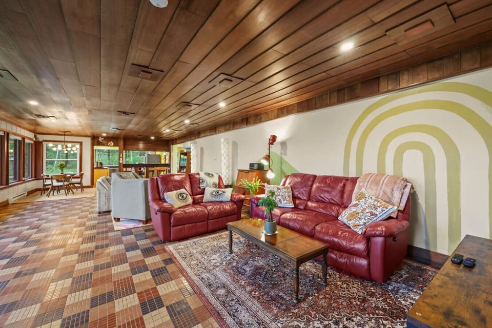 Spacious open-concept living area featuring warm wood-paneled ceilings, patterned tile flooring, and red leather sofas arranged around a coffee table, with dining space visible in the background.