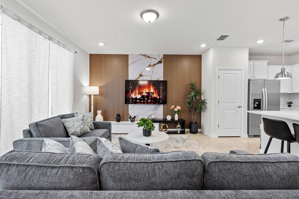 Enjoy a cozy living area with soft gray furnishings and inviting pillows, ideal for relaxation. The sleek electric fireplace adds warmth, while large windows bathe the space in natural light, creating a welcoming atmosphere.