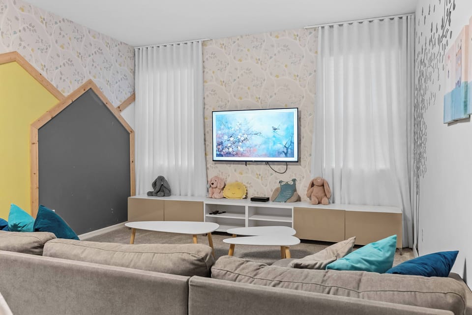 Unwind in the cozy living room with a plush sectional sofa and vibrant teal cushions. The soft pastel wall design and natural light create a warm atmosphere, while the sleek white coffee table is perfect for snacks or game nights.
