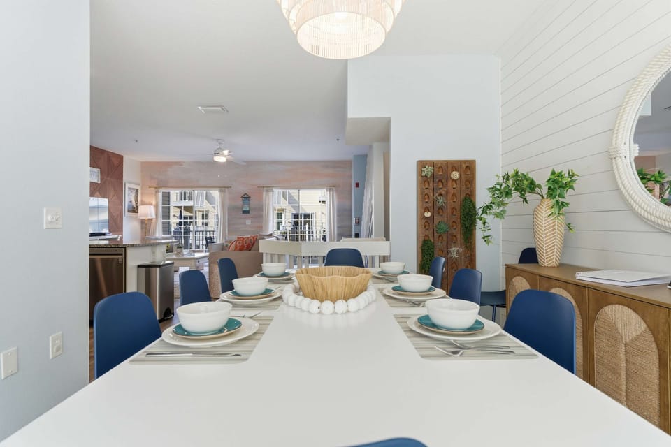 Bright and inviting dining area with a sleek white table, navy chairs, and coastal accents. Open to the living space, it’s perfect for family dinners, game nights, or gathering after a day at the beach.