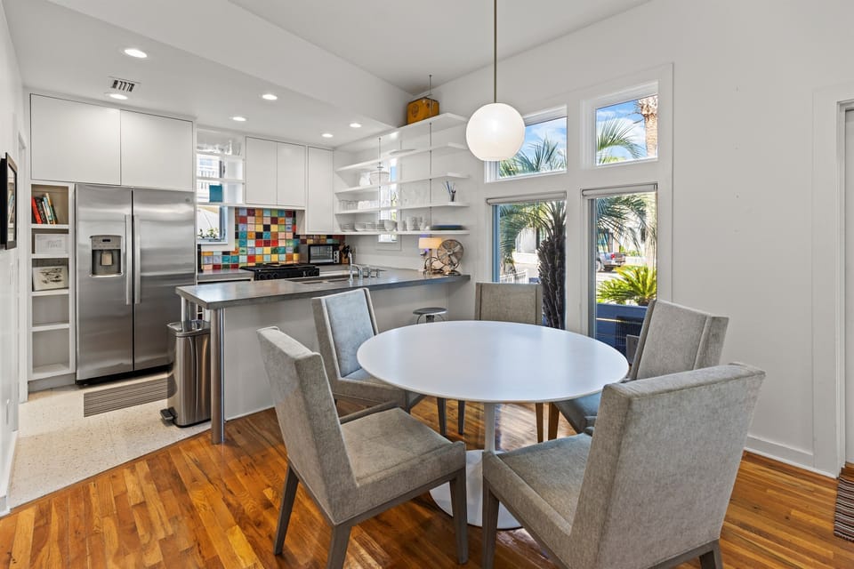 Your modern kitchen flows seamlessly into the dining space, where natural light streams through windows showcasing tropical palms outside.