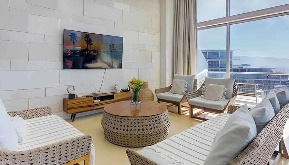 Bright living area w/ sofa seating, wall-mounted TV & floor-to-ceiling windows framing ocean views
