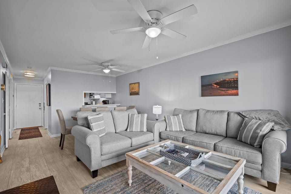 Oak Island, NC living room with gray sofas, wall TV, ceiling fan, sliding doors, ocean-view deck, coastal décor, and bright beach house style.